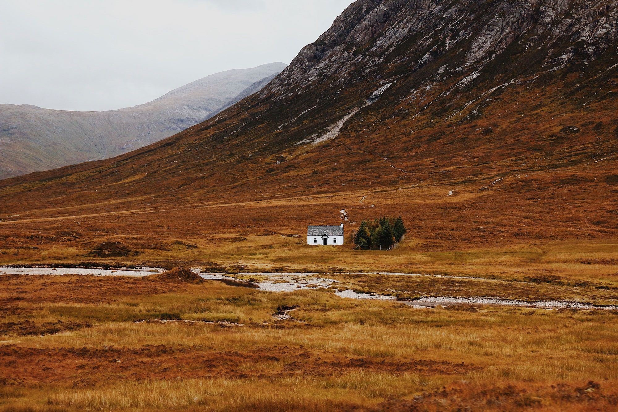 Impressive photograph of a small farm house in the isolated Glencoe valley. It’s Autumn, a small stream flows in front of the farm and a vast mountain can be seen in the background.