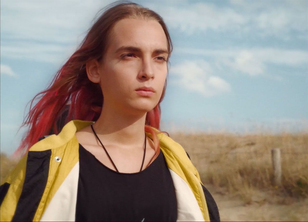 Still from feature film Lola vers la mer. Medium close up of stunning transgender teen actress Mya Bollaers looking into the horizon. There’s blue sky above her and a few clouds. In the background there are yellow turfs of dry beach grass. 