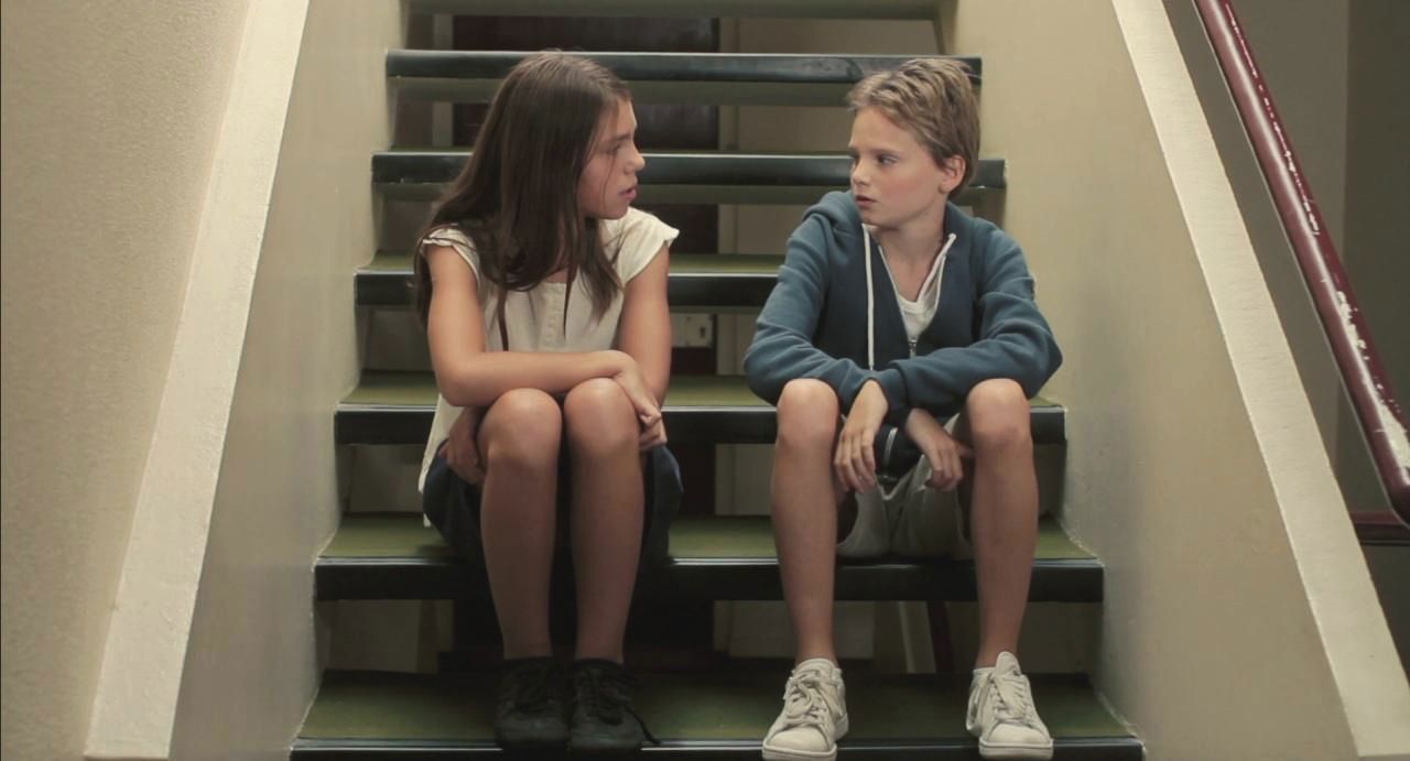 Still from the feature film Tomboy. Two kids sit and chat on a staircase. A feminine looking girl and a girl who passes as a boy.