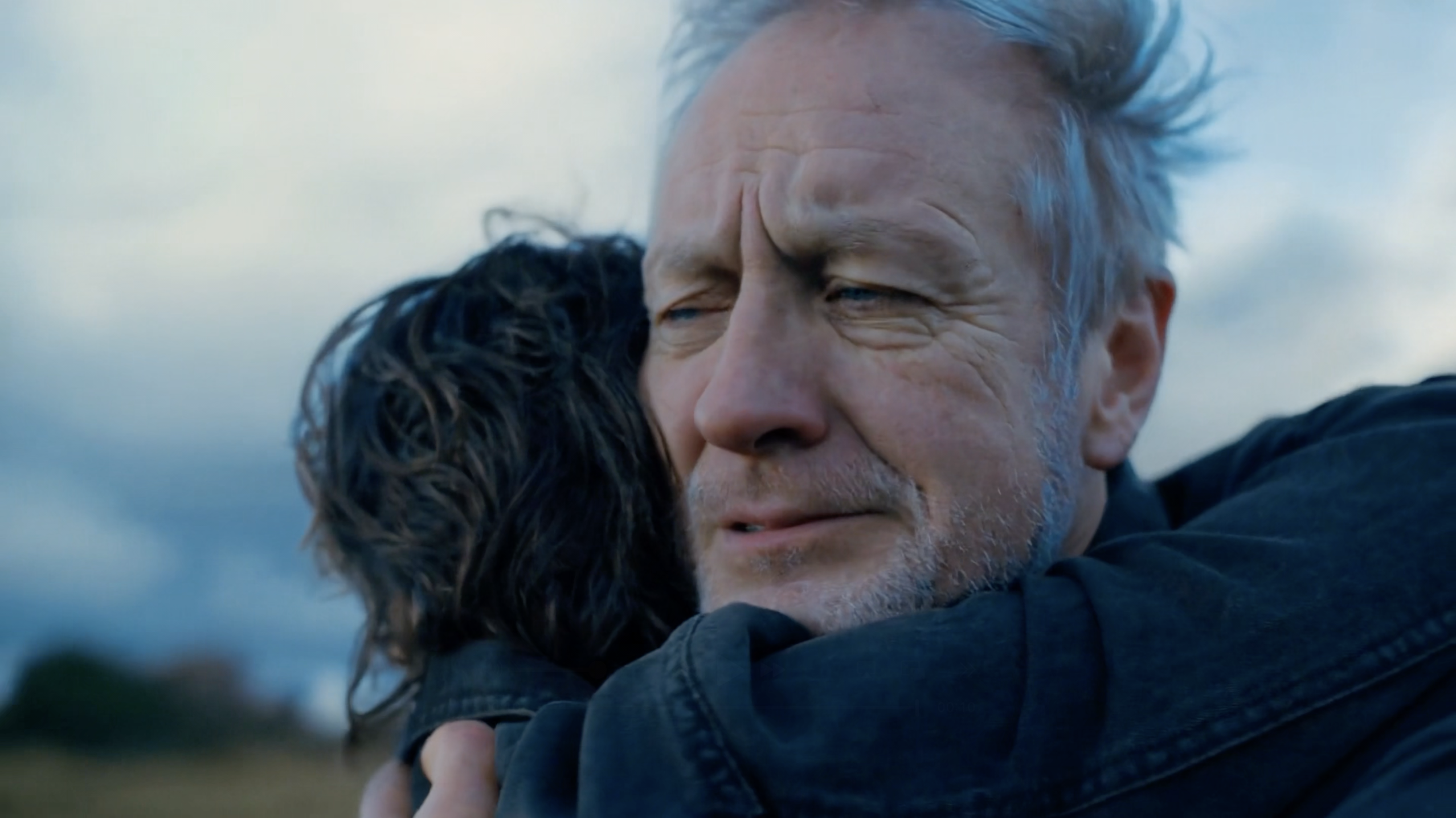 An older man with grey hair and a weathered face holds a younger person tightly in an embrace. His eyes are closed, his expression strained and emotional, as he clings to them against an open landscape under a cloudy sky.